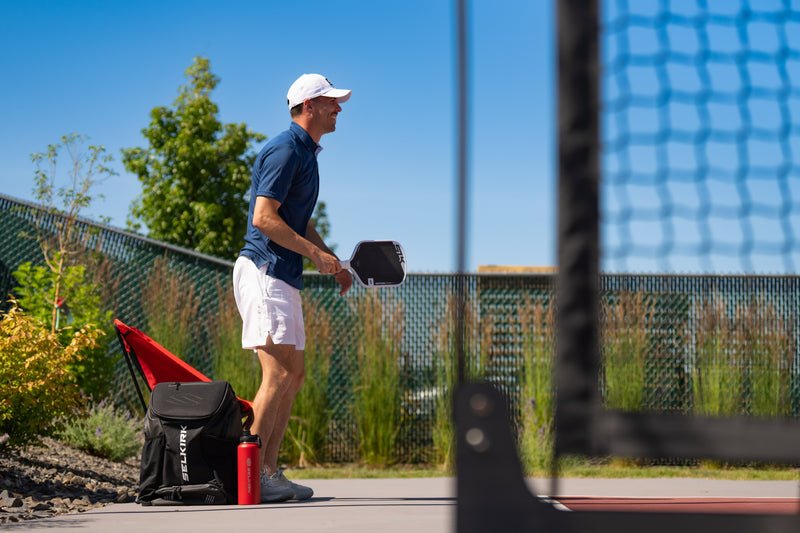 Man in white cap and blue shirt holding a pickleball paddle on an outdoor court; backpack labeled 'APOLLO' and red bottle at his feet.
