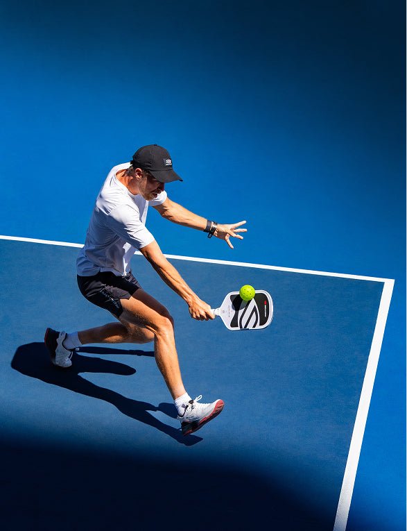 High-angle action shot of a male pickleball player in a white shirt and black shorts lunging to hit a yellow ball with a black and white Apollo Axis pickleball paddle on a blue court.
