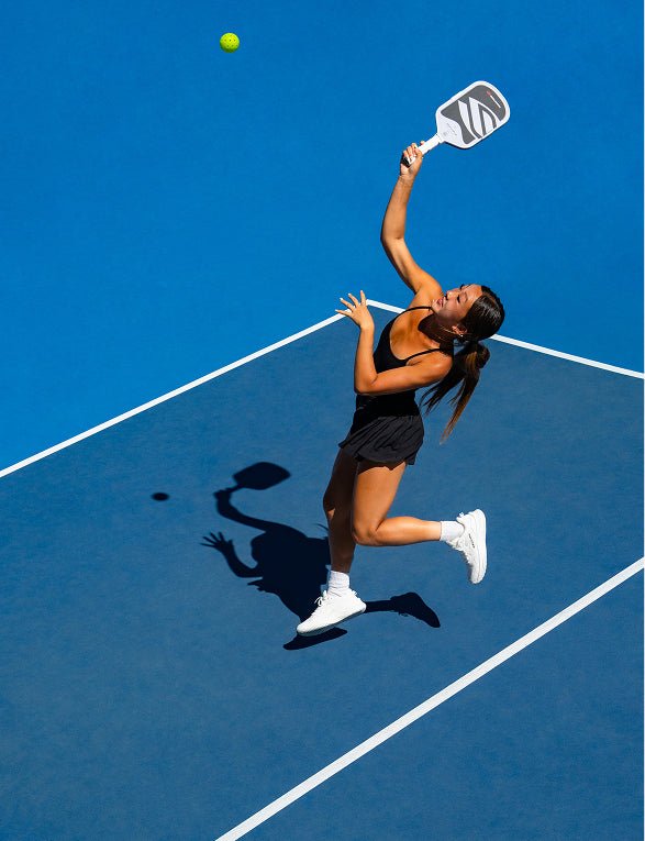 An aerial view of a female athlete executing an overhead smash shot with a white and black Apollo Axis pickleball paddle on a blue court.